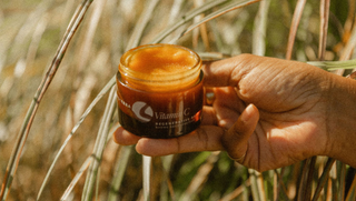a woman's hand holds Apoterra Vitamin C Regenerative Balm in an outdoor field setting near some spring reeds