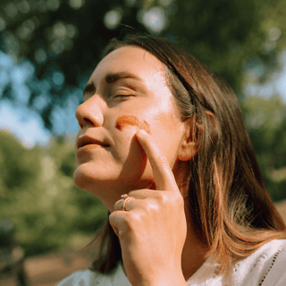 Close-up of a woman applying a Apoterra Hibiscus Mud onto her cheek in bright natural light.