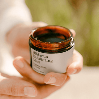 A woman's hands holding an open amber glass jar of Apoterra Hibiscus Exfoliating Mud against a soft, blurred outdoor background.