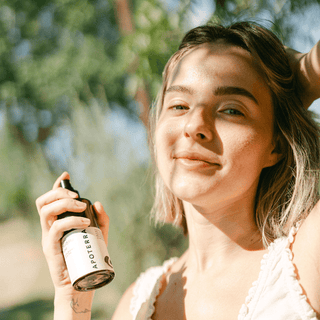 A woman outdoors holding an Apoterra facial mist bottle. The photo is brightly lit with natural sunlight and a soft blurred green background.