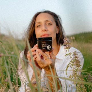 Model holding a jar of apoterra's hibiscus exfoliating mud in a field of flowers