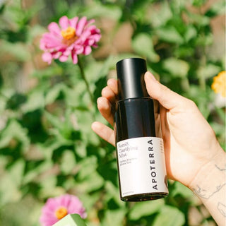 A hand holding a bottle of Apoterra Neroli Clarifying Mist against a soft-focus background of green foliage and pink flowers.