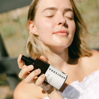 A woman holding a bottle of Apoterra Neroli Clarifying Mist outdoors with a soft focus, wearing a white sleeveless top.
