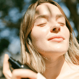 A close-up lifestyle shot of a woman outdoors with her eyes closed, holding an Apoterra facial mist bottle under warm sunlight.