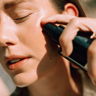 Close-up of a woman applying a Apoterra Vetiver Oil Cleanser from a dark pump bottle onto her cheek in bright natural light.