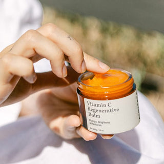 A close-up of a person's hands holding an open jar of Vitamin C Regenerative Balm, showing the bright orange balm inside. The jar has a white label with black text and is held against a soft-focus outdoor background.