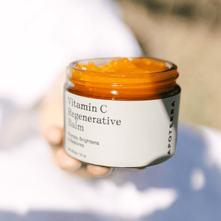 A hand holding an open amber glass jar of Apoterra Vitamin C Regenerative Balm, showing the bright orange balm inside against a soft, blurred white and beige background.