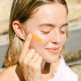 A close-up lifestyle shot of a woman outdoors applying bright orange swatch of vitamin c regenerative balm to her cheek with her finger.