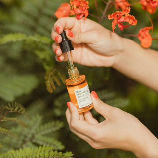 Dropper being removed from Apoterra Rose Nourishing Facial Oil bottle in front of vibrant coral flowers that match the color of the oil in the bottle
