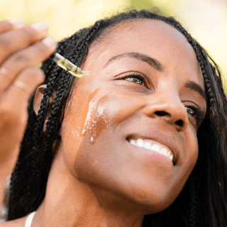 A close up of a woman with braided hair applying Green Tea Clarifying Facial Oil to her cheek in a natural outdoor setting