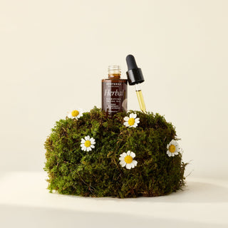 Herbal Clarifying Facial Oil by Apoterra sits on a moss block with flowers. The dropper top is off and shows the herbal oil. 