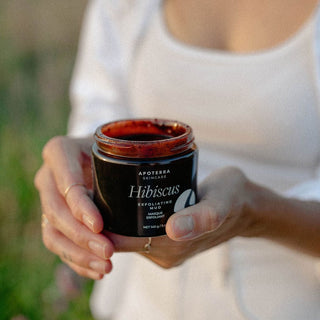 Hibiscus Exfoliating Mud Mask by Apoterra being held by model outside in a field showing rich color and texture of the mask in the jar