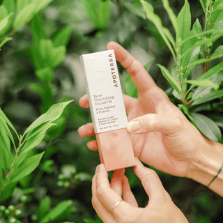 A woman's hands holding Apoterra Rose Facial Oil within its box in an outdoor setting surrounded by green leaves