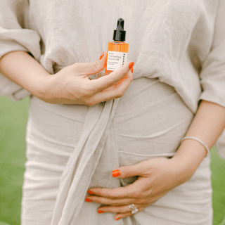 A pregnant woman's abdomen holding Apoterra Rose Facial Oil in one hand and caressing her belly with the other hand in an outdoor natural setting
