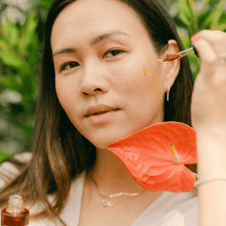 A woman holding a dropper of Apoterra Sea Berry Balancing Oil to her cheek in an outdoor setting surrounded by tropical plants