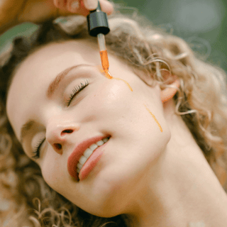 a blonde woman applying a dropper of Apoterra Sea Berry Balancing Oil to her cheek in a natural outdoor setting
