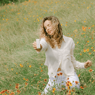 A woman kneeling in a grassy field of orange and yellow wildflowers, holding up an Apoterra facial oil box with one hand while wearing a cream-colored button-up shirt and white pants.